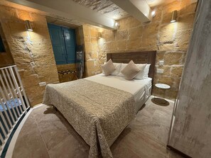 A bedroom with a stone wall, a bed with patterned bedding, a small round table, and a blue door. - Valletta City Suites Barrakka + Knight's Rest (Valletta)