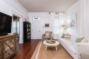 A living room with a television, a bookshelf, a door with a sign, a sofa, a coffee table, and a lamp. - Sunny, Updated Cottage in Hillcrest / Mid-Term Rental House (Little Rock)