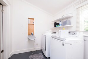 A laundry room with a washing machine, dryer, ironing board, and shelves with storage items. - Sunny, Updated Cottage in Hillcrest / Mid-Term Rental House (Little Rock)