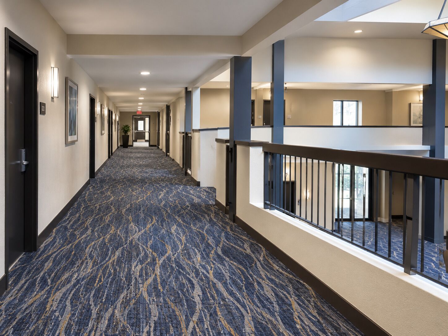 Un couloir d’hôtel avec un tapis à motifs, des portes et un garde-corps donnant sur un niveau inférieur.