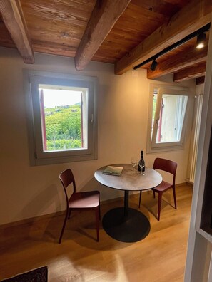 A room with a round table, two chairs, a bottle, and two wine glasses. There are two windows with views of greenery.