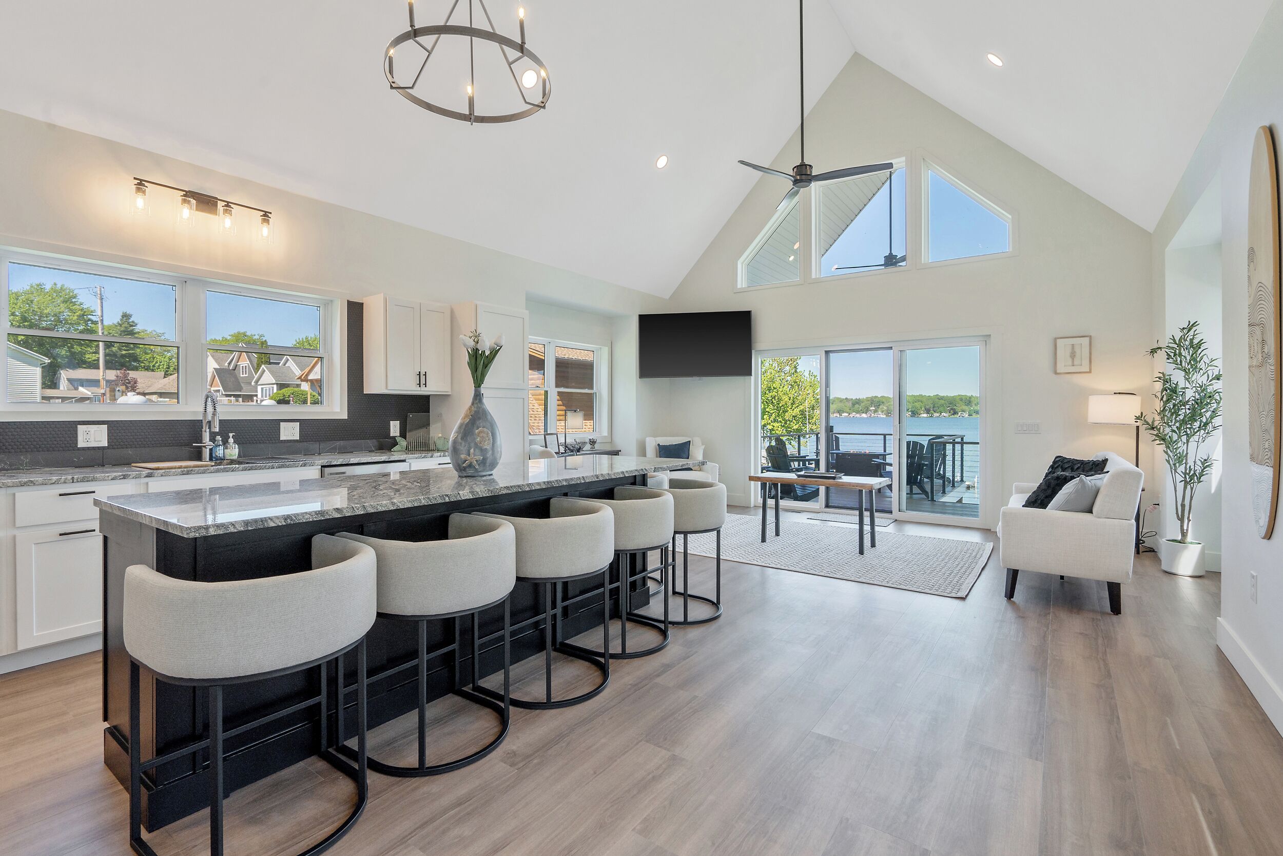 A modern kitchen with a central island, bar stools, a dining area with a view, and a living space with a large window overlooking the outdoors.