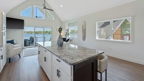 A spacious kitchen with a large island, a dining area with a view, and a living room with a high ceiling and large windows. - Shoreline Retreat - New Modern Luxury Waterfront Escape -Dock & Water Toys (Sterling)