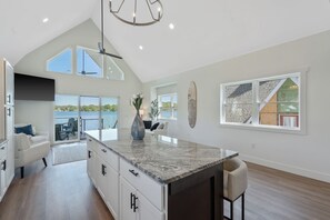 A spacious kitchen with a large island, a dining area with a view, and a living room with a high ceiling and large windows. - Shoreline Retreat - New Modern Luxury Waterfront Escape -Dock & Water Toys (Sterling)