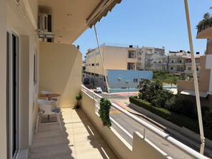 A balcony with a table and chairs, overlooking a street with buildings and a blue structure. - Chrysa Apartment (Iraklio)
