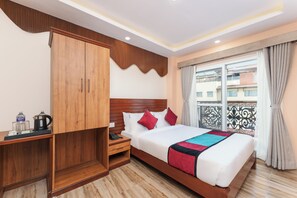 A hotel room with a wooden wardrobe, a bed with white bedding and colorful pillows, a bedside table with a kettle and glasses, and a window with curtains. - Aarohan Hotel Thamel (Kathmandu)
