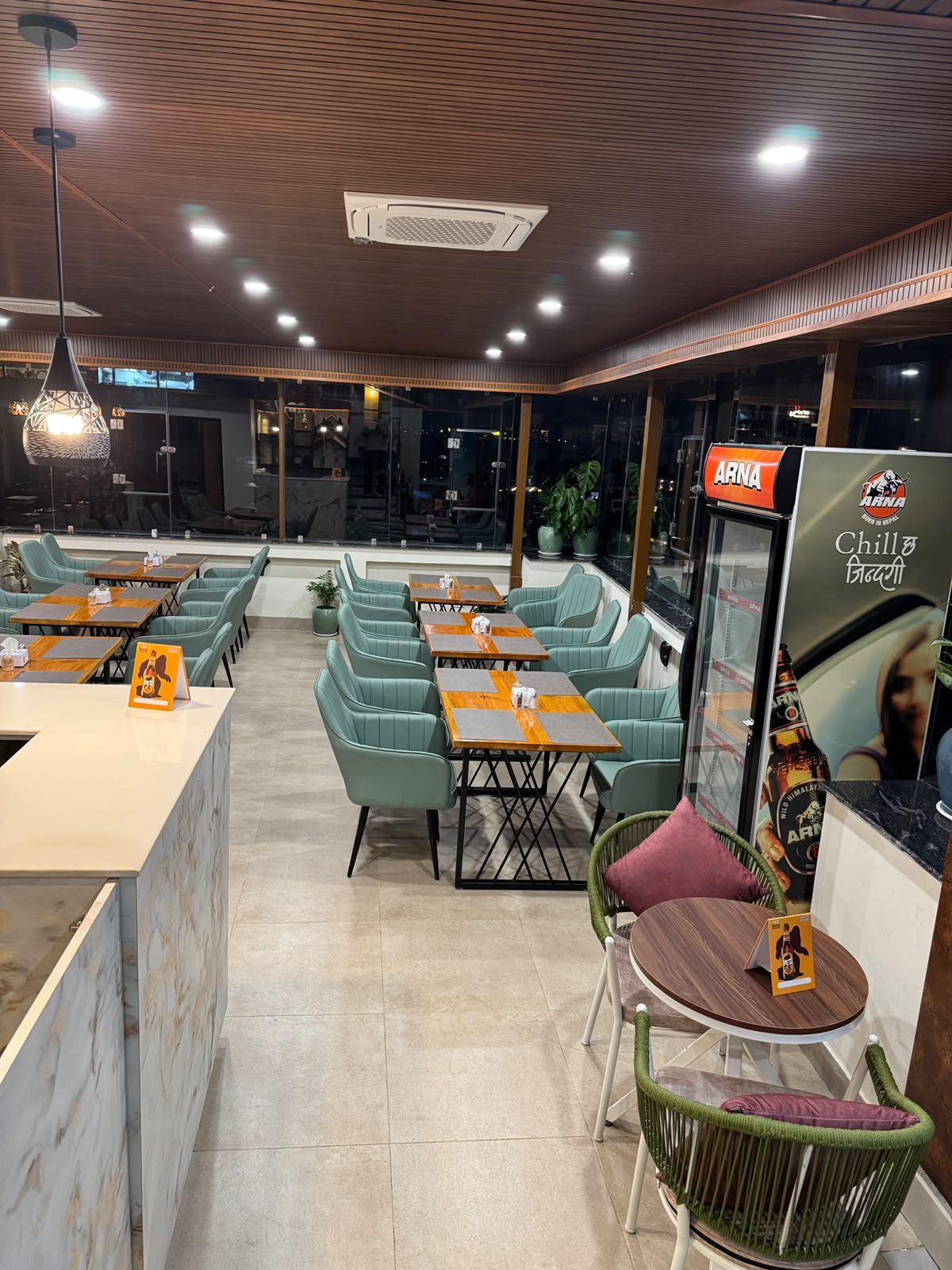 Un interior de restaurante con disposición de mesas, una barra y un refrigerador con la marca.