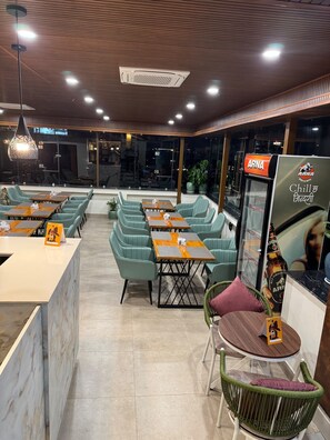 Un interior de restaurante con disposición de mesas, una barra y un refrigerador con la marca.
