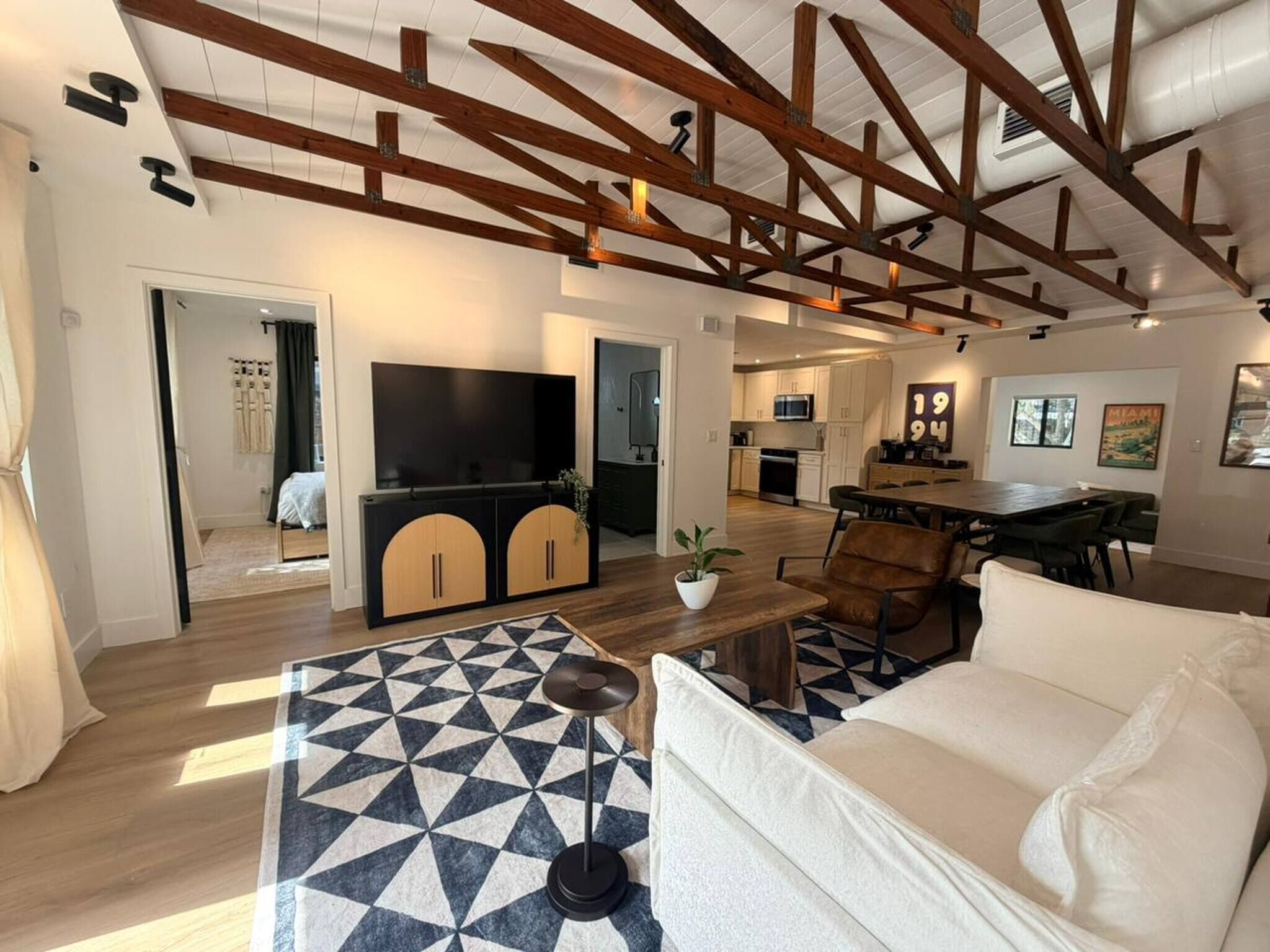 Uma sala de estar espaçosa com uma televisão grande, um sofá e uma mesa de centro. Cozinha visível ao fundo, com armários e uma área de refeições.