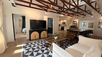 Uma sala de estar espaçosa com uma televisão grande, um sofá e uma mesa de centro. Cozinha visível ao fundo, com armários e uma área de refeições.