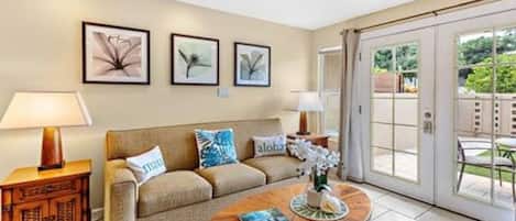 A living room with a sofa, coffee table, and lamps. There are framed pictures on the wall and a sliding glass door leading to an outdoor area.