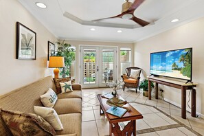 A living room with a sofa, television, ceiling fan, and a view of the outdoors through a glass door. - Steps to Maui's Charley Young Beach Condo Apt 3 (Kihei)