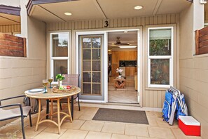 A covered patio area with a table set for two, a wicker chair, and a red cooler. - Steps to Maui's Charley Young Beach Condo Apt 3 (Kihei)