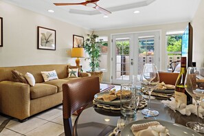 A living room with a sofa, a dining table set for a meal, and a view through a glass door. - Steps to Maui's Charley Young Beach Condo Apt 3 (Kihei)