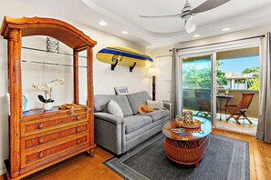 A living room with a wooden cabinet, a grey sofa, a round coffee table, and a surfboard on the wall. - Steps to Maui's Charley Young Beach Condo Apt 5 (Kihei)