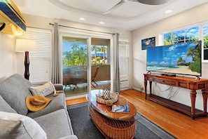 A living room with a sofa, a television on a wooden stand, a window with a view of palm trees, and a ceiling fan. - Steps to Maui's Charley Young Beach Condo Apt 5 (Kihei)