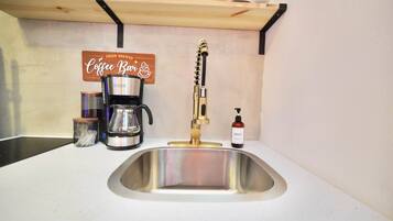 Une cuisine équipée d’un évier en acier inoxydable, d’une machine à café, d’un distributeur de savon et d’une étagère ornée d’une enseigne représentant une tasse de café.