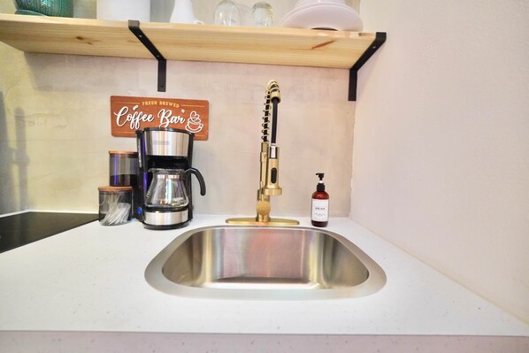 A kitchen with a stainless steel sink, a coffee maker, a soap dispenser, and a shelf with a coffee sign.