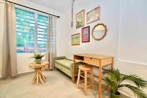 Ein Zimmer mit einem Bett, einem Schreibtisch, einem Hocker, einem kleinen Tisch mit Pflanze und Bildern an der Wand.
