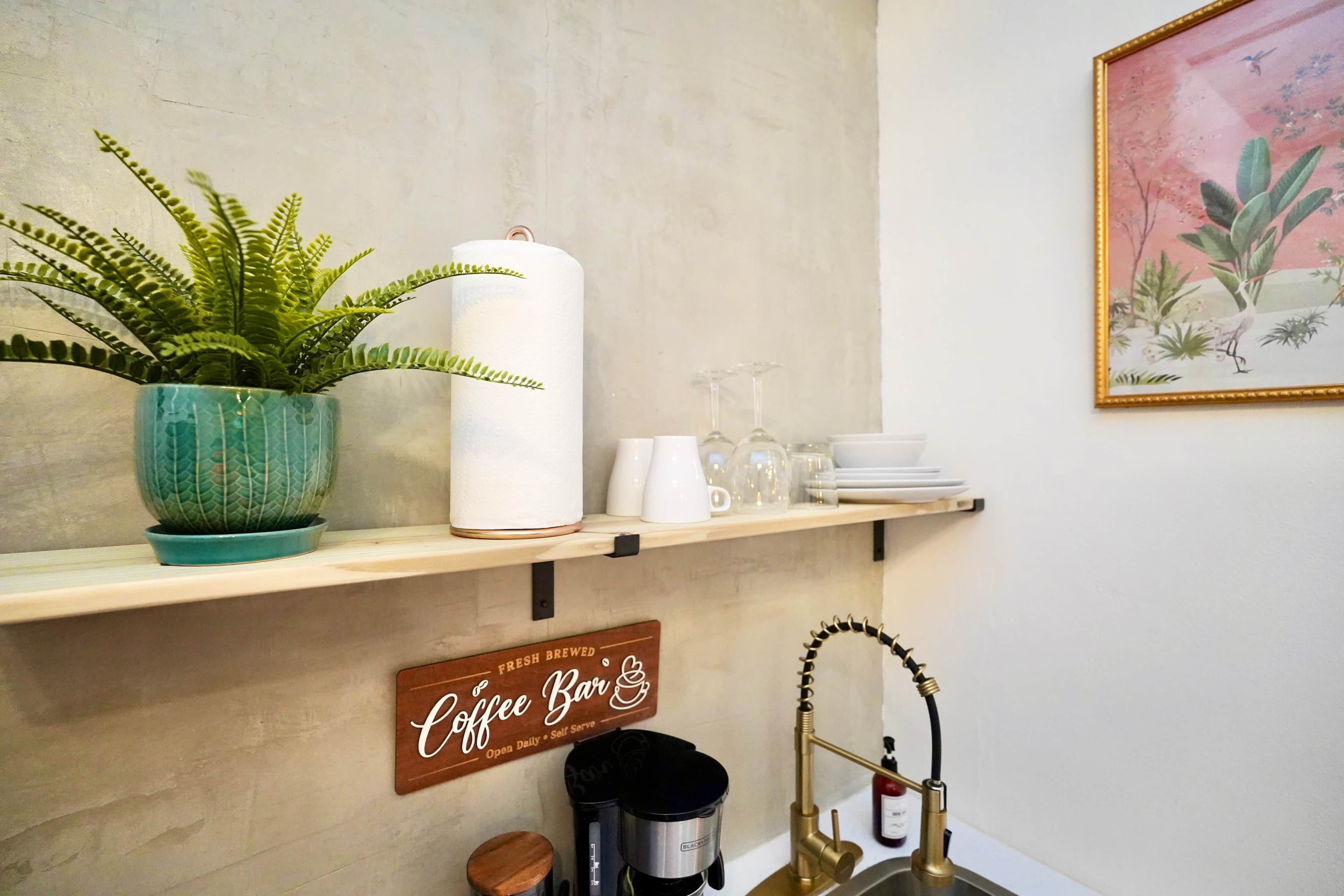 Un espacio de cocina con un letrero de barra de café, un helecho en maceta, una vela blanca y una cafetera.