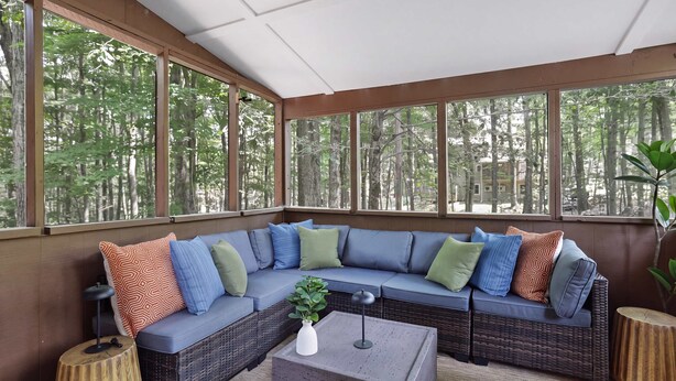 A sunroom with a sectional sofa, a small table, and a view of a forest through large windows. - Pocono Lake Escape | Hot Tub, Games & Outdoor Fun (Monroe County)