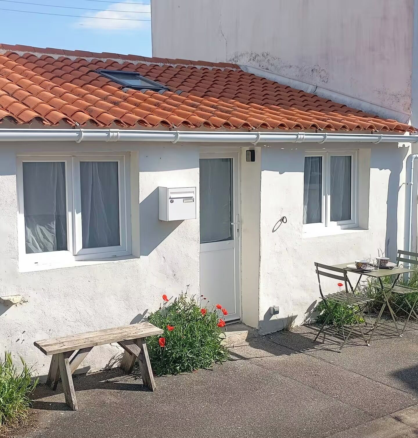 A small, single-story building with a tiled roof, a white exterior, and a door with a window. There is a bench and a table with chairs outside.