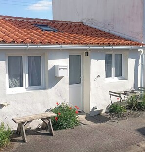 A small, single-story building with a tiled roof, a white exterior, and a door with a window. There is a bench and a table with chairs outside. - Charming Little House Near the Beach and Forest (La Tranche-sur-Mer)