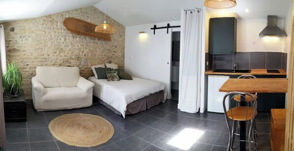 A compact room with a bed, a chair, a small kitchenette, and a stone wall.