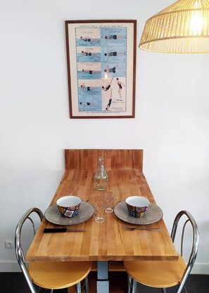 A dining table with two chairs, a framed instructional poster, and a hanging light fixture. - Charming Little House Near the Beach and Forest (La Tranche-sur-Mer)