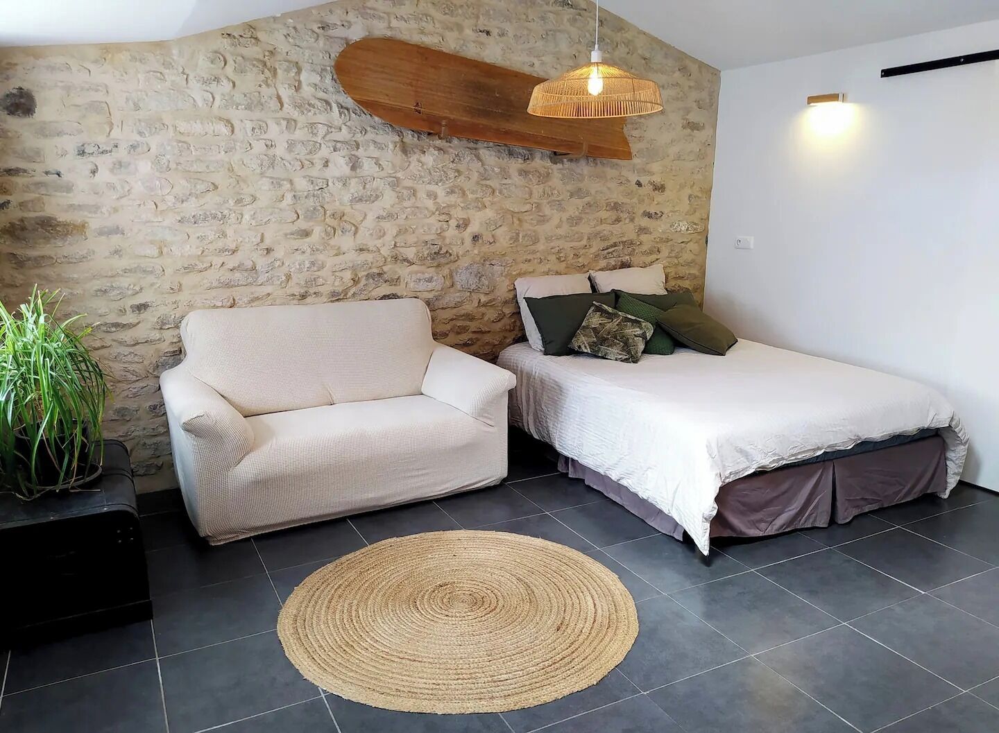 A bedroom with a stone wall, a sofa, a bed with pillows, a hanging light, and a woven rug.