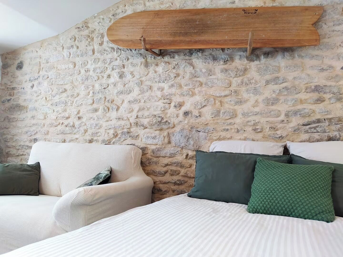 A bedroom with a stone wall, a bed with white bedding, and a wooden surfboard mounted on the wall.