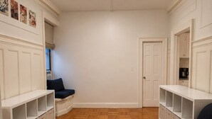 Huoneessa on kalanruotokuviollinen puulattia, valkoiset, seinään integroidut hyllyt, tummanvärinen sohva ja ovi, joka johtaa toiseen huoneeseen.