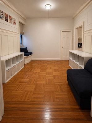 Ein Zimmer mit einem Fischgrätparkett, weißen Einbauschränken, einem dunkelgrauen Sofa und einer Tür zu einem anderen Raum.