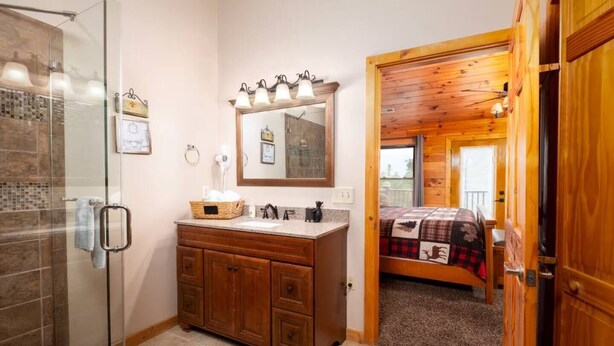 A bathroom with a glass shower enclosure, wooden vanity, and a bed with a plaid blanket. - Mountain Views & Hot Tub | 1 Mi to Ober (Gatlinburg)