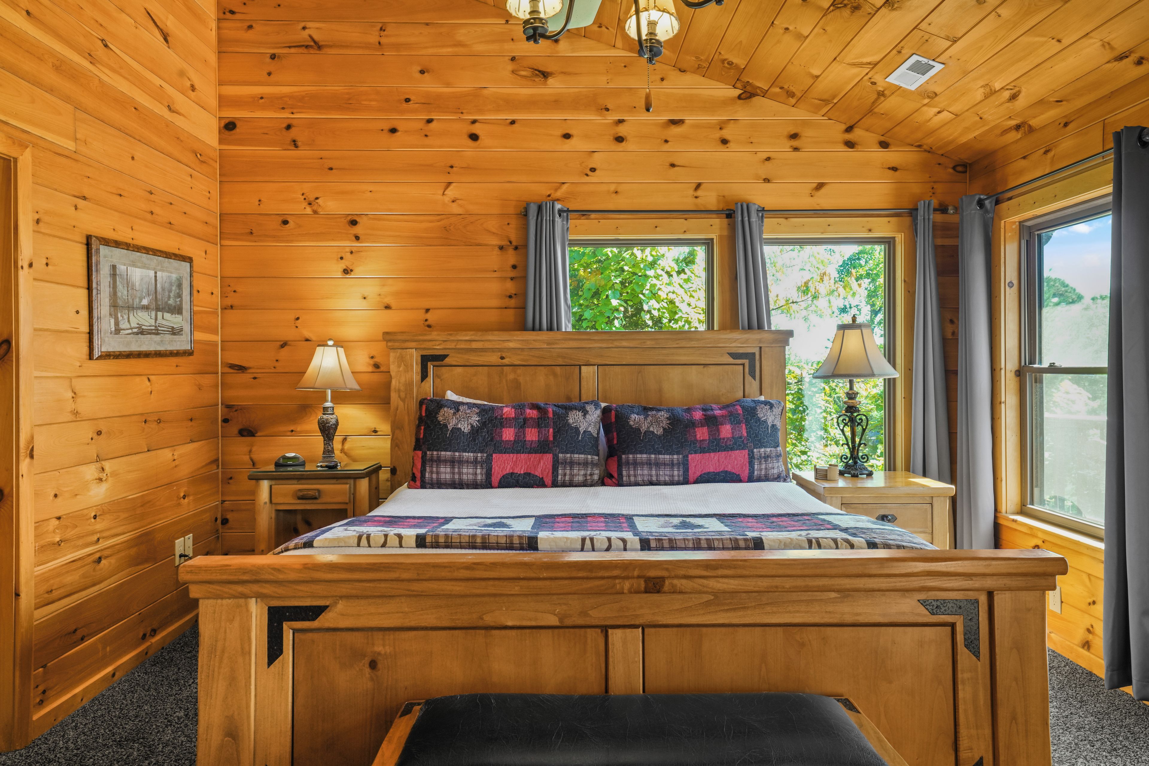 Quarto de cabana de madeira com cama, mesas de cabeceira, abajur, janela com cortinas e um quadro na parede.