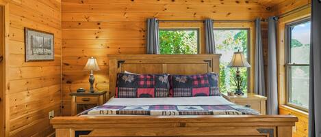 Quarto de cabana de madeira com cama, mesas de cabeceira, abajur, janela com cortinas e um quadro na parede.