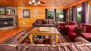A cozy cabin interior with a fireplace, wooden walls, a ceiling fan, and a large window. - Mountain Views & Hot Tub | 1 Mi to Ober (Gatlinburg)
