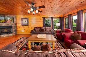 A cozy cabin interior with a fireplace, wooden walls, a ceiling fan, and a large window. - Mountain Views & Hot Tub | 1 Mi to Ober (Gatlinburg)