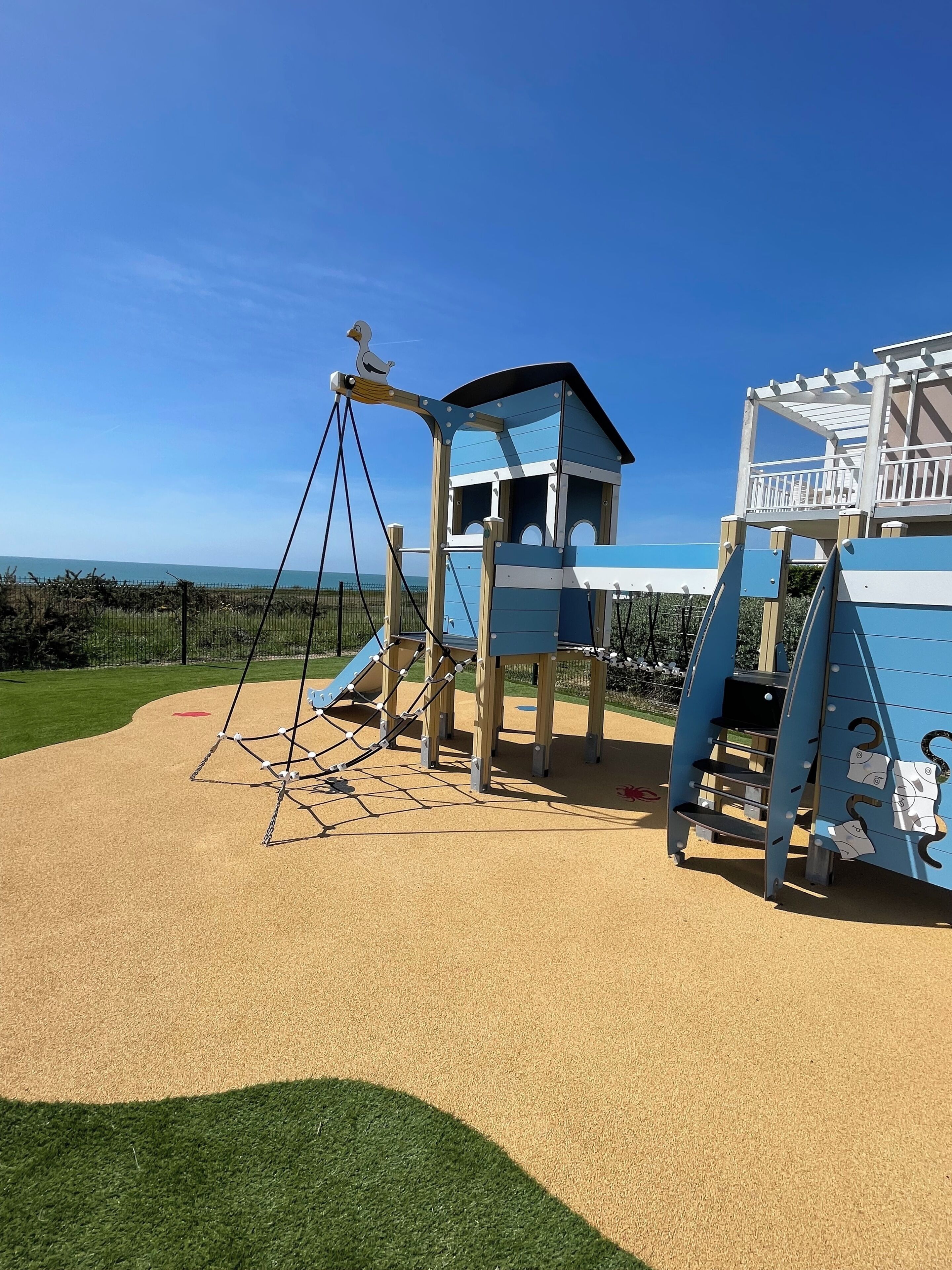 A playground with a climbing structure and a slide, surrounded by a sandy area and a grassy field.