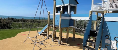 A playground with a climbing structure and a slide, surrounded by a sandy area and a grassy field.