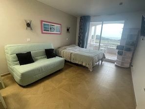 A room with a light blue sofa, a bed with a patterned bedspread, a small wall-mounted clock, and a view of the outdoors through a sliding glass door. - ☀️ Getaway with a sea view • Swimming Pools • Wild Coast (Les Sables-d'Olonne)