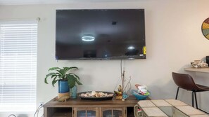 Ein Wohnzimmer mit Fernseher an der Wand, einem Esstisch, einem Topfpflanzen, dekorativen Muscheln und einer kleinen Statue.