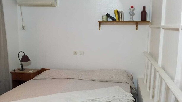 En enkelt seng med hvid og beige dyne, et natbord med lampe, samt en reol med bøger og pynteting.