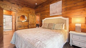 A wooden-paneled room with a large bed, a round mirror, and a bedside table with a lamp. - Hillside Retreat, Modern 2BR Cabin | Hot Tub, Pool Table & Fire Pit (Sevierville)