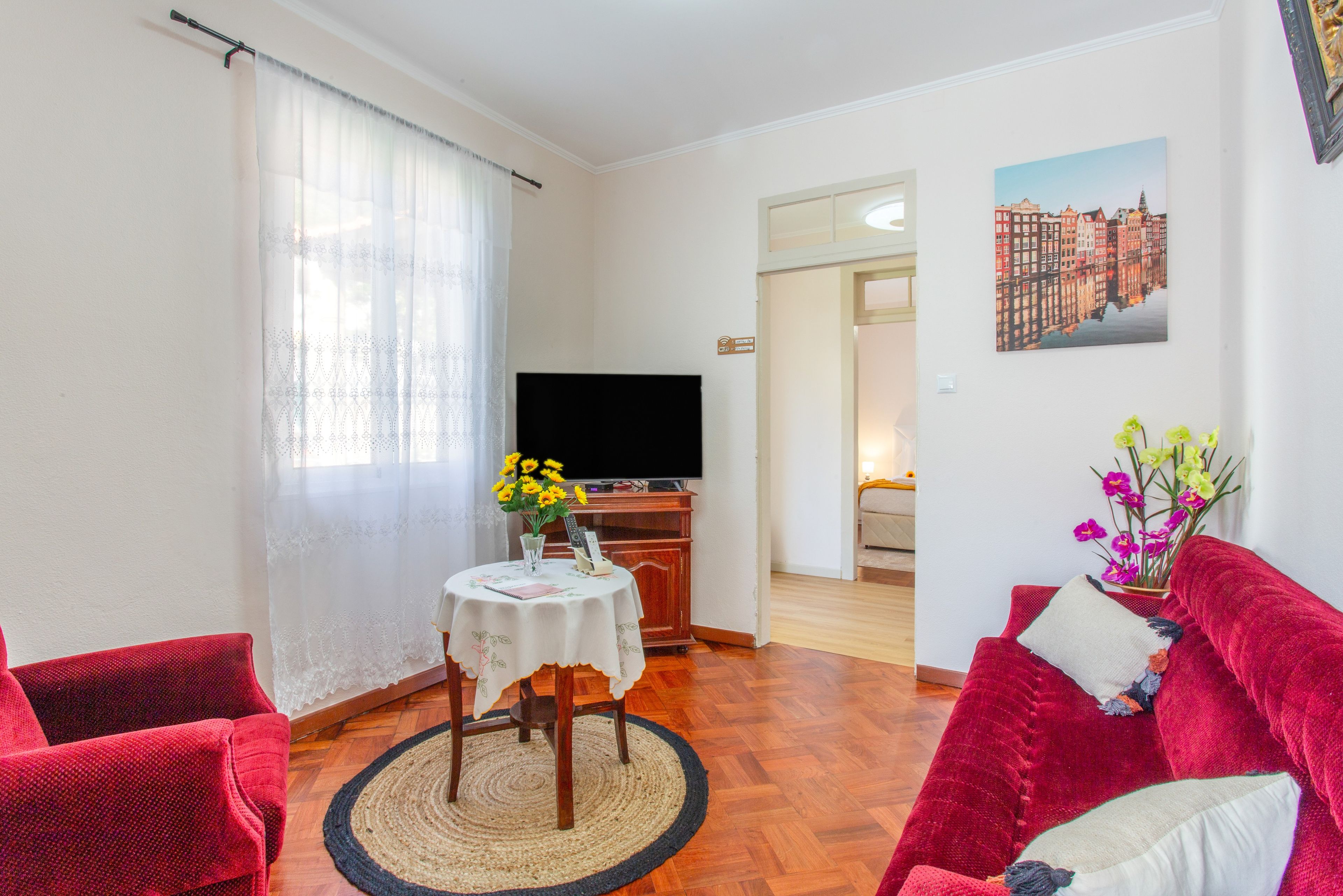 Ein Wohnzimmer mit Fernseher, einem Tisch mit Blumenvase, einem roten Sofa und einem Bild an der Wand.