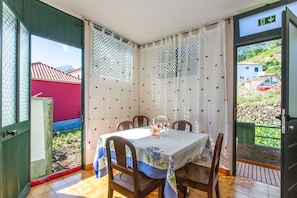 Un coin repas avec une table dressée pour deux personnes, des chaises, une fenêtre avec des rideaux et une vue sur un bâtiment aux toits rouges à l’extérieur.