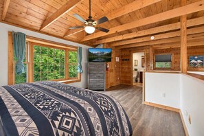 Interior d'una cabana amb un llit, un ventilador de sostre, una finestra amb cortines, un bany i un quadre a la paret.
