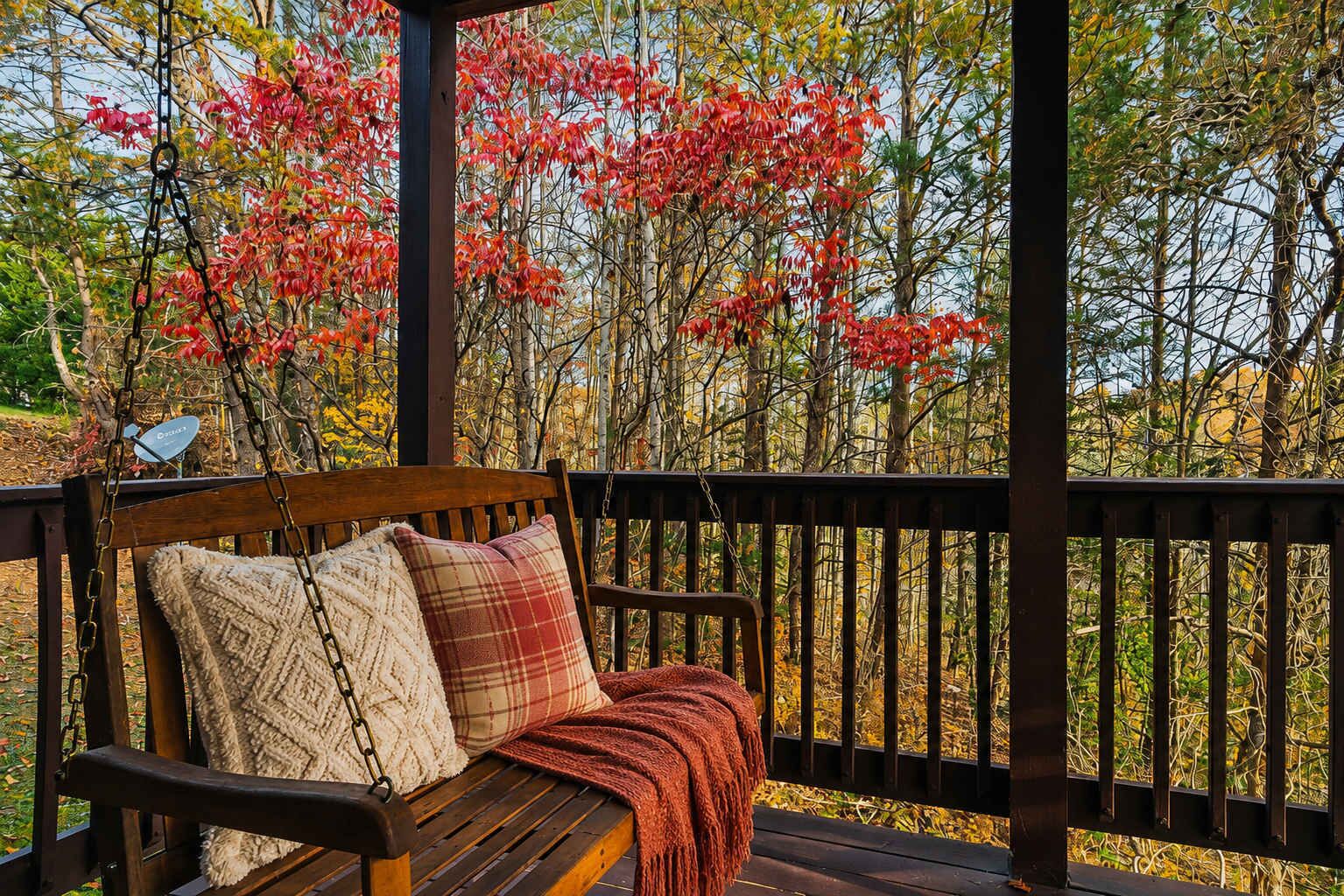 A wooden bench with a plaid cushion and a throw blanket, under a wooden pergola with a view of trees with red leaves.