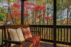 Un banc en bois agrémenté d’un coussin à carreaux et d’une couverture, placé sous une pergola en bois, d’où l’on peut admirer des arbres aux feuilles rouges.