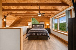 A bedroom with a sloped ceiling, a bed with patterned bedding, bedside lamps, and a view of greenery through a window. - Mountain Escape - Hot Tub - Game Room - Sleeps 8 (Pigeon Forge)